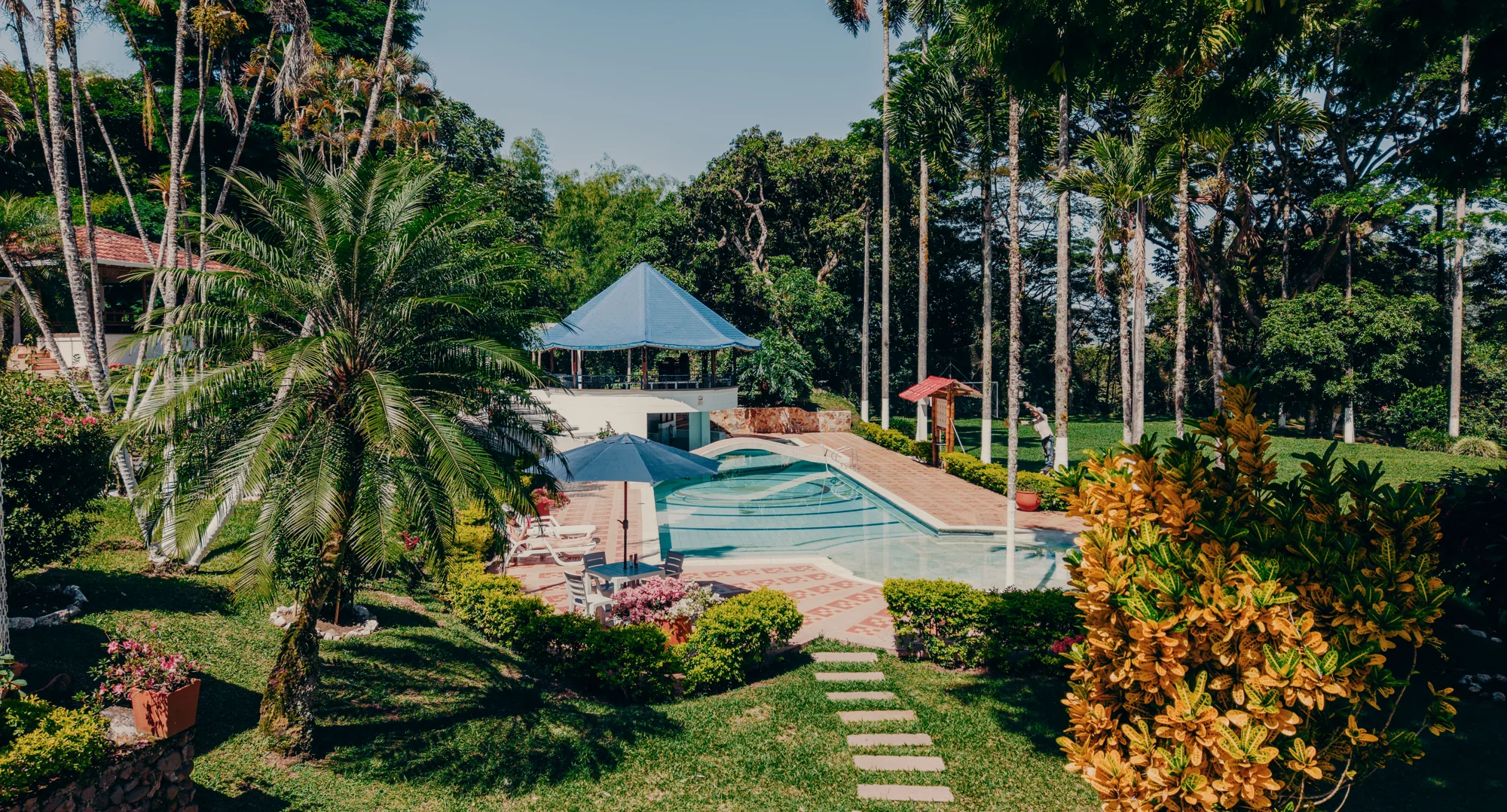 La piscina y el kiosco entre las dos casas, rodeados de palmeras y jardines